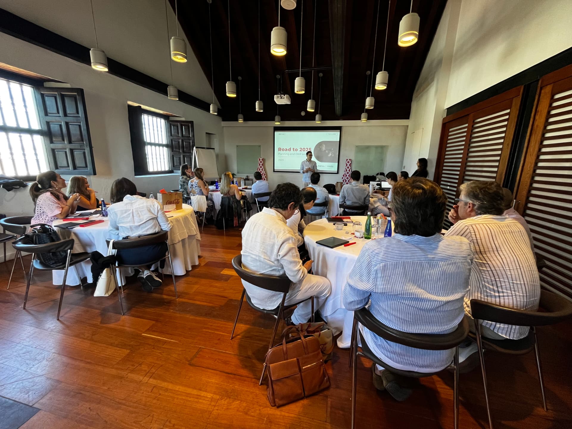 Participantes sentados em mesas redondas com notas e dispositivos, assistindo a uma apresentação intitulada Caminho para 2025